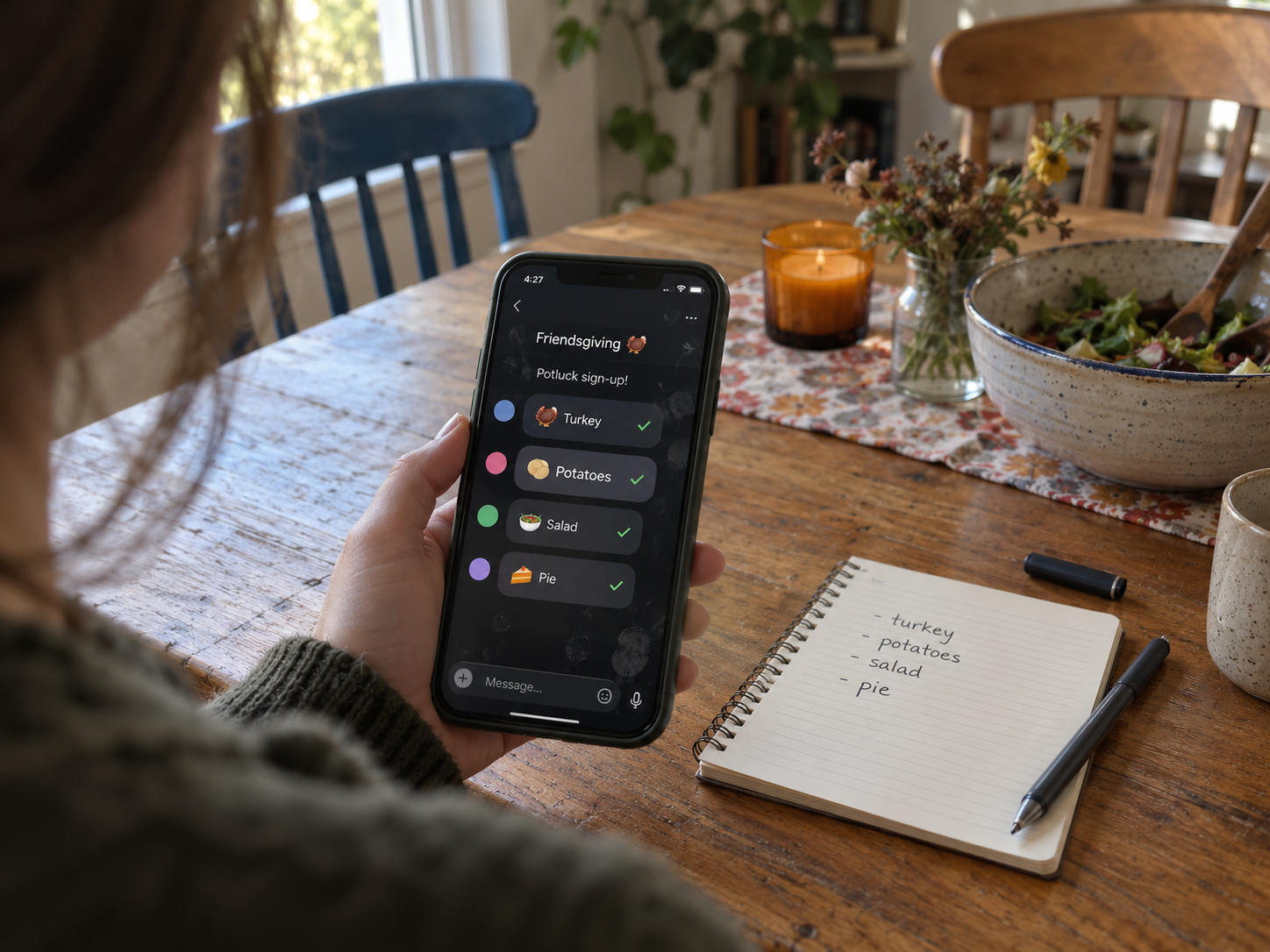 Phone showing a Friendsgiving potluck group chat beside a handwritten list of dishes on a dining table